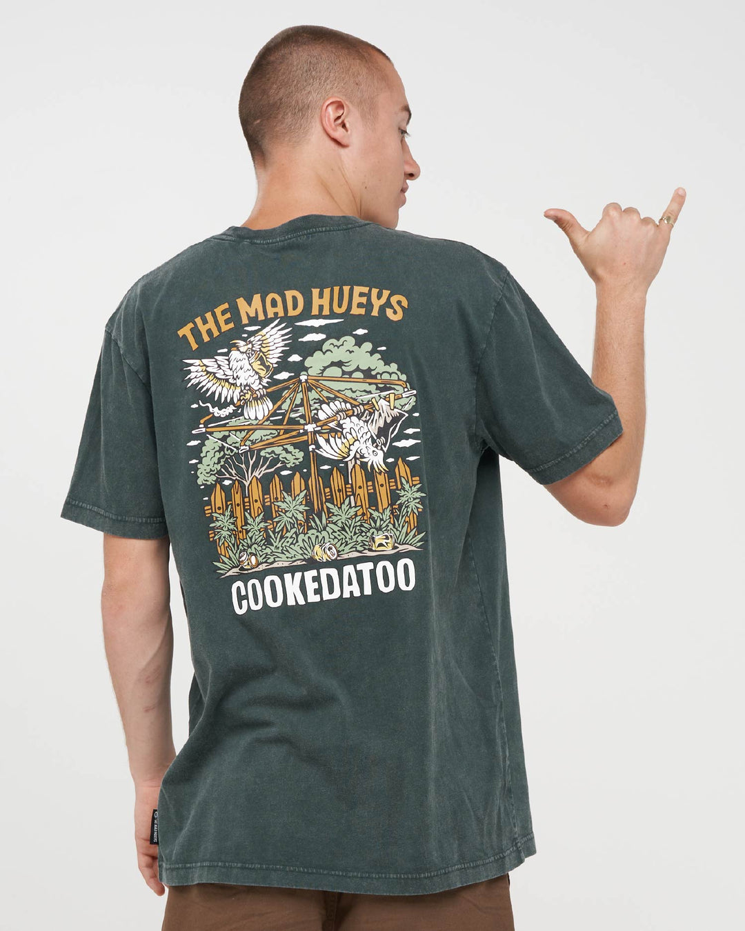 A man wearing a dark washed green graphic t-shirt, viewed from the back. The shirt features a large graphic print with eagles, a fence, and the text &